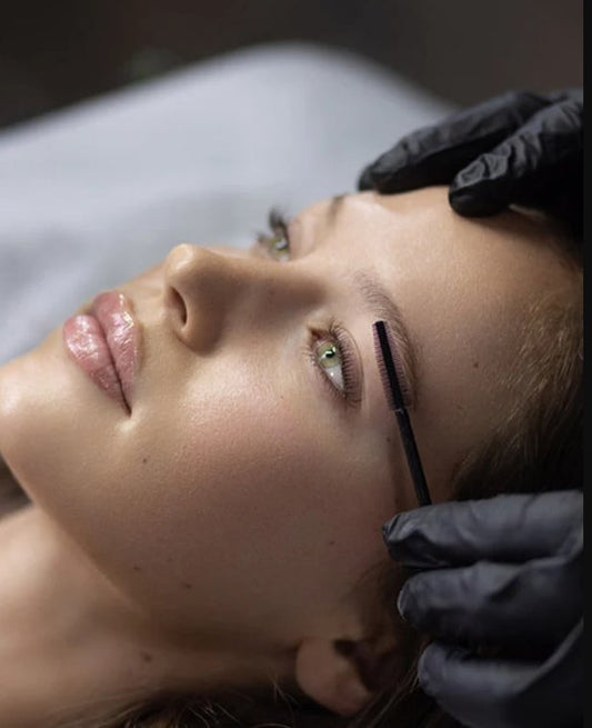 Close-up of a person receiving an eyebrow treatment as a gloved technician brushes the brow hairs with a small wand.